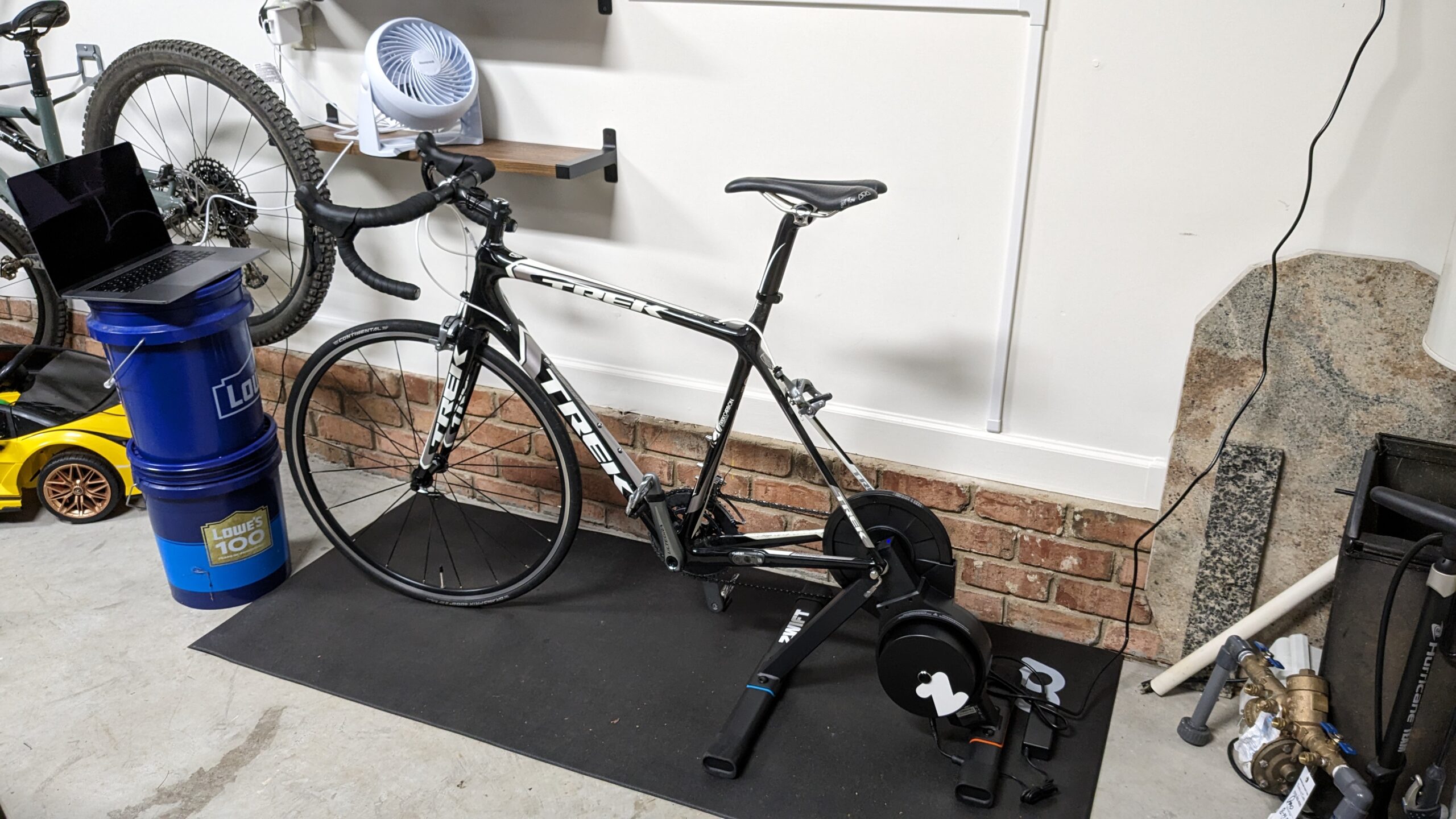 Photo of my bike mounted on the bike trainer in my garage. There's a black mat underneath, shelves on the wall with a fan on one of the shelves, and two 5 gallon Lowe's buckets stacked with a laptop on top.
